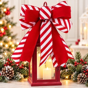 Op de afbeelding: Een rode metalen lantaarn met drie witte stompkaarsen, bekroond met een grote decoratieve strik met een rood-wit diagonaal gestreept patroon. De lantaarn heeft een rechthoekig frame en een handvat aan de bovenkant, waar de strik aan vastzit.