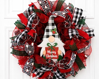 Puede incluir: Una corona navideña decorativa con malla roja y verde oscuro, cintas de cuadros negros y blancos, y un recorte central de un gnomo. El gnomo lleva un gorro de cuadros y sostiene una caja de regalo roja con el texto Merry Christmas en blanco.