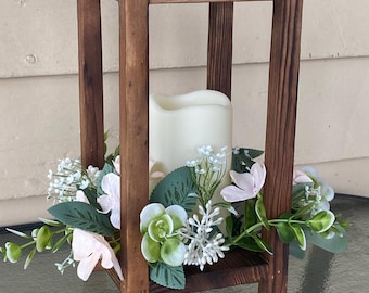 May include: A wooden candle lantern with a square frame. The lantern holds a white candle surrounded by a floral arrangement of pink and white flowers, green leaves, and white sprigs. The lantern is made of stained wood.