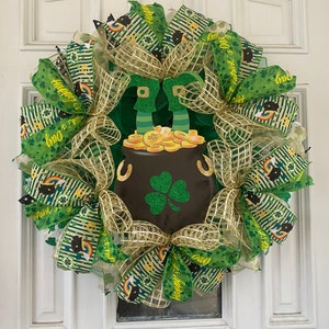 Pode incluir: Uma coroa verde, dourada e preta do Dia de São Patrício com um pote de ouro e pernas de duende. A coroa é decorada com fita e malha verde e dourada. A fita tem o texto "Feliz Dia de São Patrício" e "Sorte dos irlandeses".