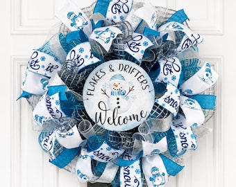 May include: A decorative wreath featuring blue, white, and silver mesh ribbons with snowflake patterns and snowman illustrations. A central circular sign displays a snowman graphic with the text Flakes & Drifters Welcome in blue script.