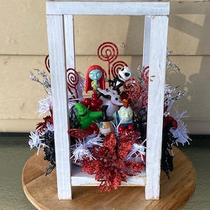 Op de afbeelding: Een wit houten lantaarnframe met een verzameling Nightmare Before Christmas-figuren, waaronder Jack Skellington, Sally, Zero, Oogie Boogie en anderen, geaccentueerd met rode glitterspiralen, rode bessen en witte klatergoudversieringen.