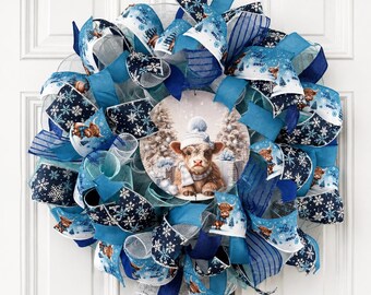 May include: A decorative wreath featuring blue and white mesh ribbons with snowflake patterns and a central circular sign depicting a Highland cow wearing a winter hat and scarf. The design incorporates various shades of blue and white fabric.