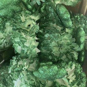 May include: Close-up of a collection of artificial flowers with a glittery green finish. The flowers have a layered petal design and are in varying shades of green. The glitter adds a festive touch to the floral arrangement.