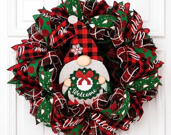 Puede incluir: Una corona decorativa con una figura central de gnomo que lleva un gorro de cuadros rojos y negros con un copo de nieve blanco. El gnomo sostiene una corona verde con un lazo rojo y el texto Welcome. La base consiste en cintas rojas, verdes y de cuadros.