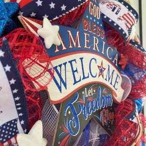 Pu&ograve; includere: Una ghirlanda patriottica rosso, bianco e blu per la porta con un cartello a forma di stella che recita "God Bless America Welcome Let Freedom Ring".