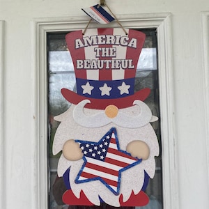 Puede incluir: Un gnomo colgador de puerta rojo, blanco y azul con una bandera estadounidense en forma de estrella en sus manos. El gnomo lleva un sombrero que dice "America the Beautiful".