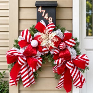 Op de afbeelding: Een decoratieve krans met groene dennentakken, rood-wit gestreepte linten, rode fluwelen linten, witte mesh accenten, een grote goudkleurige glitter kerstster en twee ronde kerstballen, gemonteerd onder een zwarte brievenbus met het nummer 4749.