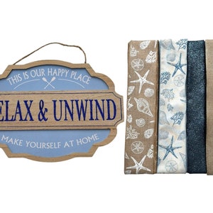Op de afbeelding: Een houten bord met de tekst "This is our happy place Relax & Unwind Make yourself at home" in blauwe letters. Het bord is versierd met twee gekruiste roeispanen en een blauwe achtergrond. Het bord wordt vergezeld door vier rollen lint met een nautisch thema.