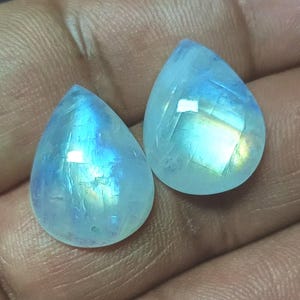 Puede incluir: Dos cabujones de piedra lunar en forma de lágrima con colores iridiscentes azules y blancos.