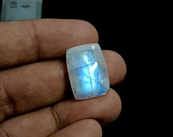 May include: A rectangular moonstone cabochon with a translucent white body and a vibrant blue adularescence glow. The gemstone is positioned next to a ruler showing a measurement of approximately 0.5 inches in width for the stone's top edge.