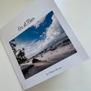 May include: A white booklet with the title "En el Mar" in black cursive writing. The cover features a photograph of a beach with blue sky, white clouds, and palm trees.