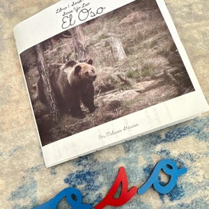 May include: A book titled "El Oso" with a photograph of a brown bear in a forest. The book is open, and the title is written in cursive. Below the book are wooden letters spelling "oso", with the "o"s in blue and the "s" in red.