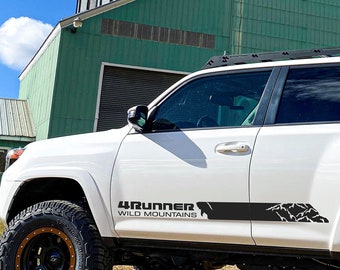 May include: White Toyota 4Runner SUV with black roof rack and black side decals that say "4Runner Wild Mountains". The vehicle is parked in front of a green building with a mural of wheat stalks.