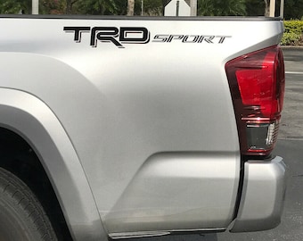 May include: A silver pickup truck with a black "TRD SPORT" decal on the side. The truck has a black bumper and a black tire.