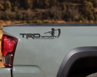 May include: A gray pickup truck with a black "TRD Sport" decal on the tailgate. The decal features a silhouette of a person holding a scythe.
