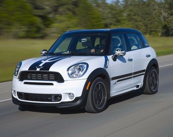 May include: A white Mini Cooper Countryman with black accents and a black roof driving on a paved road. The car has black racing stripes on the hood and sides.