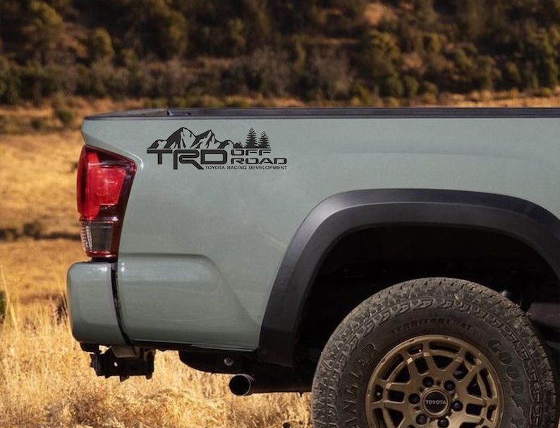 May include: A black TRD Off Road decal on the side of a light green truck bed. The decal features mountain and tree graphics with the text "TRD OFF ROAD TOYOTA RACING DEVELOPMENT". The truck has a black tire and a red tail light.