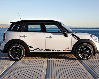 May include: A white Mini Cooper S Countryman with black accents and a checkered racing stripe along the side. The car is parked on a gray concrete surface with a body of water in the background.