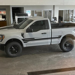 May include: A white Ford F-150 pickup truck with black accents and black wheels. The truck is parked in a showroom with a gray concrete floor.