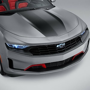 Peut inclure: Une voiture de sport convertible argentée avec une bande noire sur le capot et des accents rouges sur le pare-chocs avant. La voiture a une calandre noire avec un logo Chevrolet chromé.