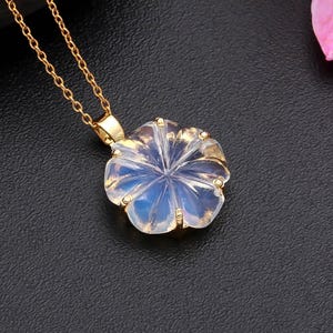 May include: A gold-colored necklace with a flower-shaped pendant. The pendant is a light blue and clear color, with a detailed floral design. The necklace is on a black surface, with a pink flower and a dark gray stone in the background.