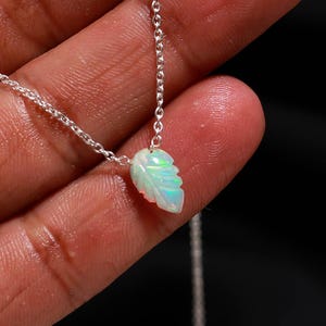 Puede incluir: Un delicado collar de cadena plateada con un pequeño colgante de ópalo tallado en forma de hoja. El ópalo muestra destellos iridiscentes de luz verde y azul sobre una base blanca translúcida, suspendido de una fina cadena de eslabones metálicos.
