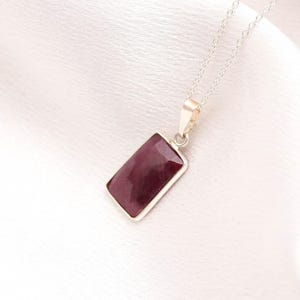 May include: A necklace featuring a rectangular, deep red gemstone pendant set in a silver-toned metal frame. The pendant hangs from a delicate, thin silver-toned chain that connects to the top of the setting with a small, simple metal bail.