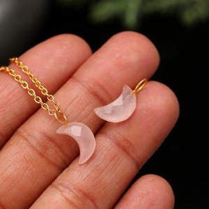 May include: A gold chain necklace with a small, faceted, pink crescent moon pendant. A matching moon charm is next to the necklace. The jewellery is displayed against a blurred background.
