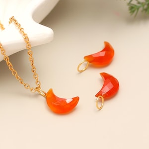 Puede incluir: Un collar de cadena dorada con un colgante de media luna y dos amuletos a juego. Los colgantes en forma de luna son de color naranja vibrante. La joyería se exhibe sobre una superficie clara.