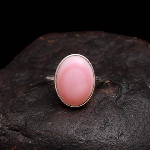 May include: A silver ring with a large, oval pink gemstone. The gemstone is set in a silver bezel and has a smooth, polished surface. The ring is displayed on a dark, textured surface, creating a contrast that highlights the ring's design.