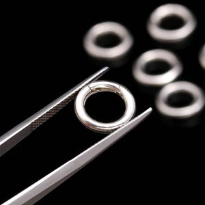 May include: Close-up of several small, shiny, silver-colored jump rings. One ring is held by a pair of tweezers, with others scattered in the background. The jump rings are circular and have a small opening. The background is black.