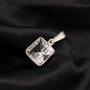 Może przedstawiać: Kwadratowa, przezroczysta kryształowa zawieszka ze srebrnym uchwytem. Kryształ jest fasetowany, odbijając światło. Zawieszka jest umieszczona na czarnym, teksturowanym tle.