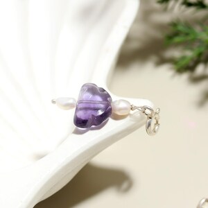 May include: A delicate piece of jewelry featuring a polished, translucent purple gemstone, flanked by two small, white pearls. The gemstone is connected to a silver clasp with a delicate chain. The jewelry is displayed on a white, seashell-shaped dish.