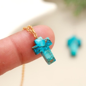 May include: A turquoise cross pendant necklace with a gold chain. The cross is a vibrant blue-green color with brown veining. The necklace is held by a finger, with a second cross pendant out of focus in the background.