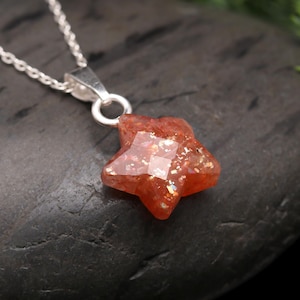 May include: A silver chain necklace featuring a star-shaped pendant. The pendant is a faceted, reddish-orange gemstone with sparkling inclusions. The necklace is displayed on a dark, textured surface with green foliage in the background.