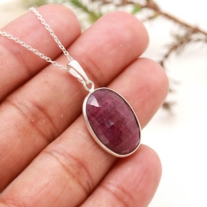 May include: A silver necklace featuring an oval-shaped, faceted deep pinkish-red gemstone pendant. The stone is set in a simple silver bezel frame and hangs from a delicate silver chain, showcasing a polished surface with natural internal textures.