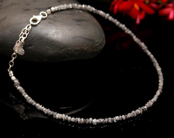 May include: A delicate silver anklet featuring a strand of small, raw, gray diamonds. The anklet is secured with a silver clasp and rests on a dark gray stone. Red flowers and a black background complete the composition.