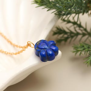 Könnte beinhalten: Eine goldene Halskette mit einem tiefblauen, blütenförmigen Anhänger. Der Anhänger besteht aus einem dunkelblauen Stein und liegt auf einer weißen, muschelförmigen Schale. Grünes Laub befindet sich im Hintergrund.