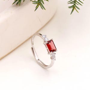 May include: A silver ring with a rectangular, deep red gemstone at the center, flanked by smaller, clear stones. The ring is set against a white background with a sprig of green foliage in the upper left corner.