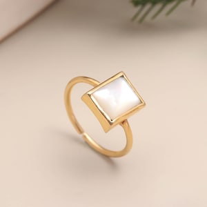 Puede incluir: Un anillo dorado con una gema blanca cuadrada. El anillo tiene una banda fina y ajustable. La gema está engastada en un bisel dorado. El anillo se muestra sobre una superficie beige claro.
