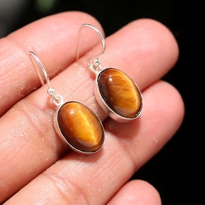 May include: A pair of oval-shaped tiger's eye earrings. Each earring features a polished, golden-brown gemstone set in a silver bezel. The earrings have a simple hook design for pierced ears, showcasing the natural beauty of the stones.