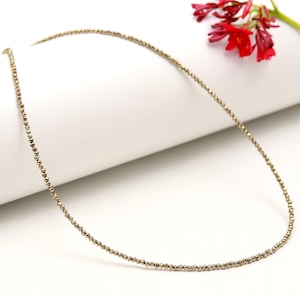 Puede incluir: Un delicado collar de oro hecho de pequeñas cuentas. El collar está sobre una superficie blanca.