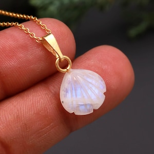 May include: A necklace featuring a gold-toned chain and a pendant carved into the shape of a small seashell. The pendant is made of a translucent, iridescent moonstone that displays a soft blue glow when light hits its polished, curved surface.