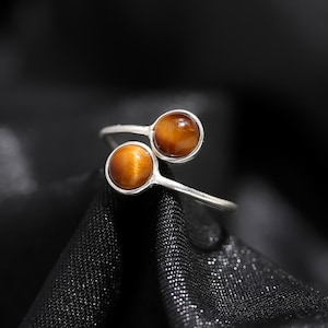 Op de afbeelding: Een zilverkleurige ring met twee gepolijste tijgeroog edelstenen. De edelstenen hebben een warme, goudbruine kleur met een chatoyance-effect. Het ringontwerp is modern en open. Diameter: ongeveer 1 cm.