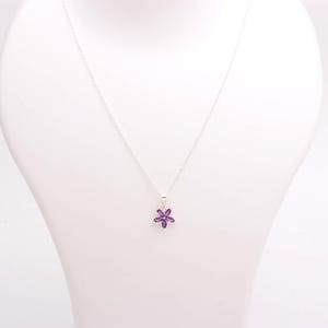 May include: A silver necklace with a small, purple flower pendant. The pendant is a six-petal flower made of small, purple gemstones. The necklace is displayed on a white mannequin.