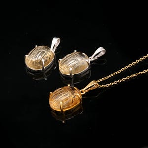 May include: Three pendants featuring scarab beetle designs. Two pendants have silver-colored bails, while the third is gold-colored and attached to a chain. The beetles are a translucent yellow color, set against a black background.