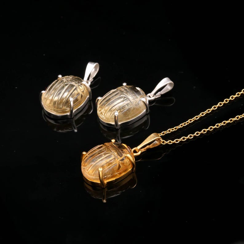 May include: Three pendants featuring scarab beetle designs. Two pendants have silver-colored bails, while the third is gold-colored and attached to a chain. The beetles are a translucent yellow color, set against a black background.