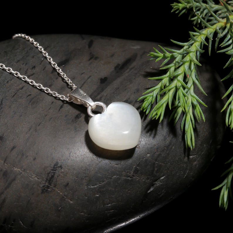 May include: A silver necklace with a heart-shaped pendant. The pendant is a smooth, opaque white stone. The chain is a delicate silver link. The necklace is displayed on a dark, textured surface with green foliage in the background.
