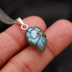 May include: A silver pendant featuring a carved blue labradorite gemstone in the shape of a leaf. The gemstone has an iridescent sheen.